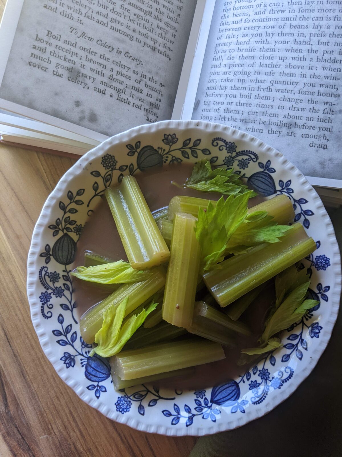 2 inch pieces of celery in a brown sauce i9n a blue and white bowl. The bowl sits on a wooden background with an open book in the background.