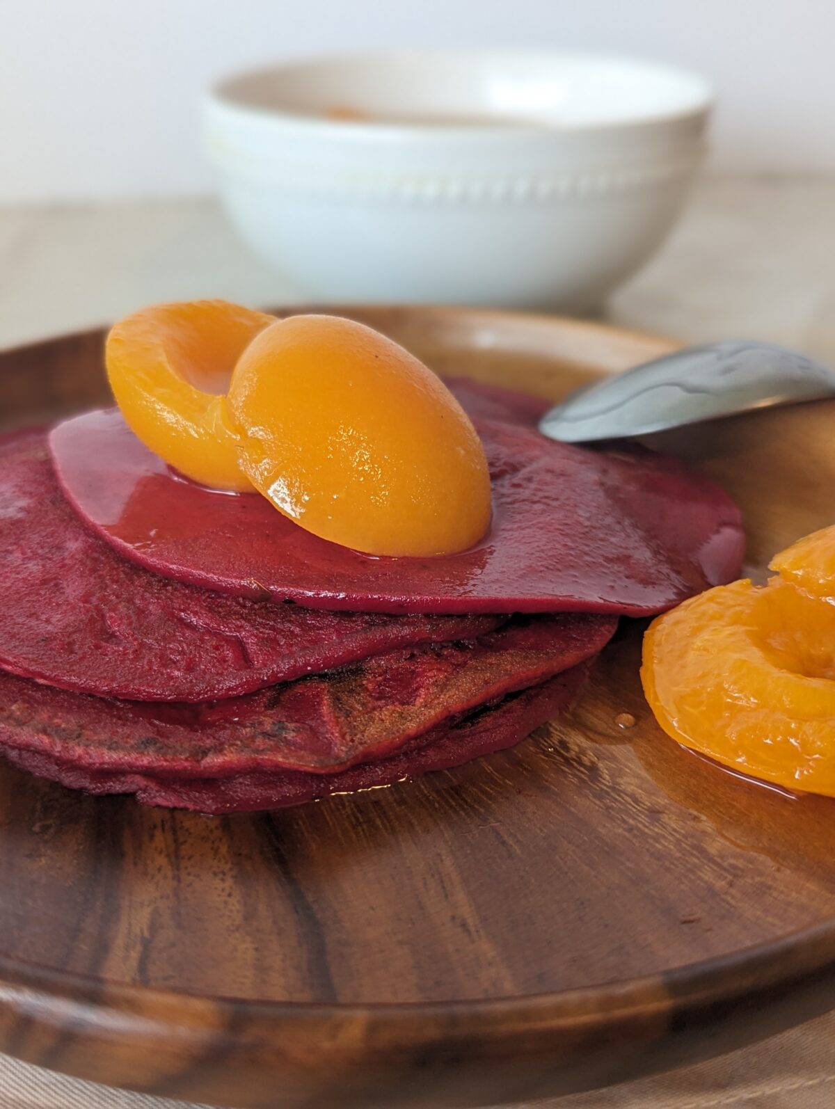 A stack of dark pink pancakes sit on a wooden plate, topped with apricots in syrup. A white bowl of apricots sits in the background, with an upturned spoon on the plate.